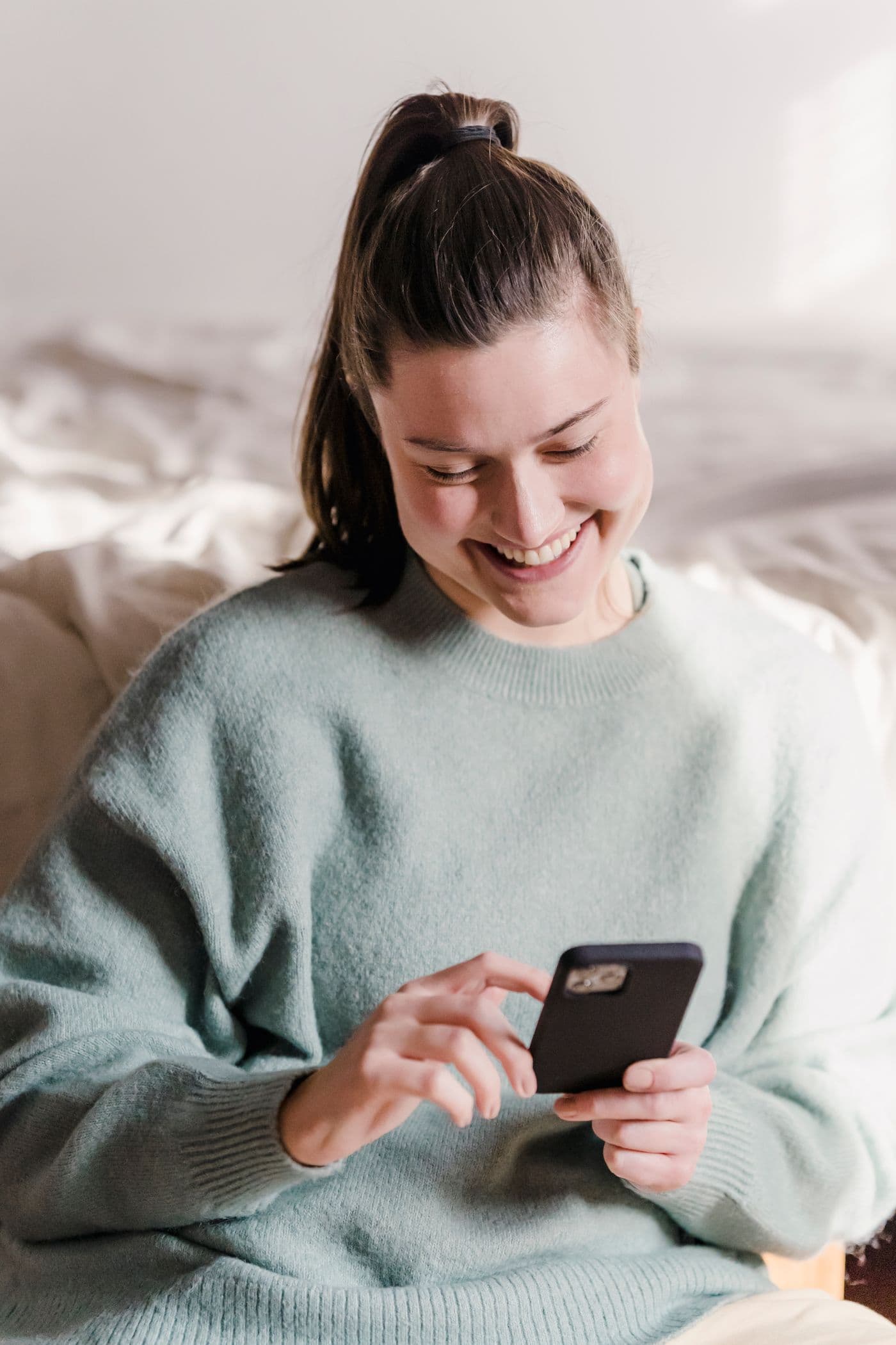 A woman sitting at home, smiling at her phone.